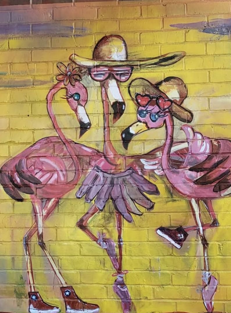 Graffiti mural of three pink flamingos wearing hats and sunglasses on a bright yellow brick wall.