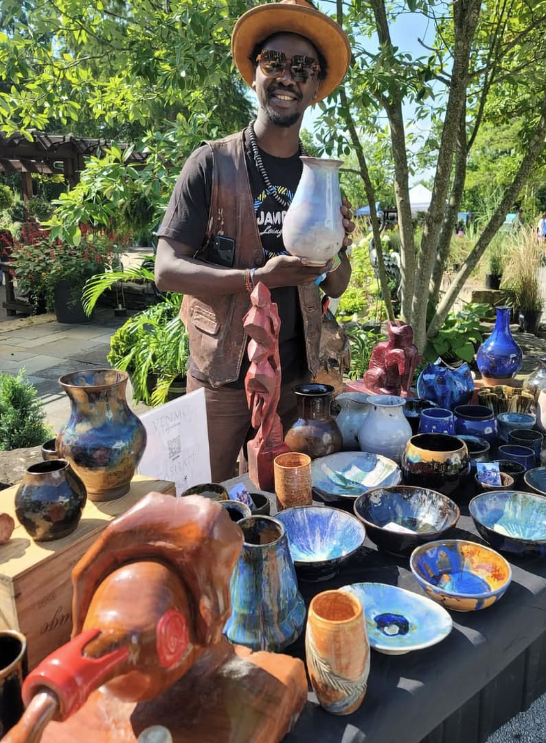 Potter wearing a hat and vest shows a ceramic market table filled with colorful bowls and vases outdoors.