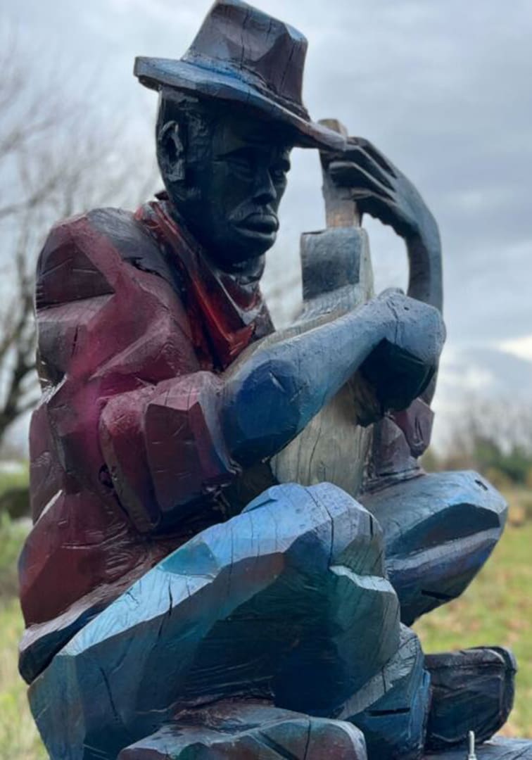 Outdoor wooden sculpture of a man wearing a hat and a red scarf, sitting and leaning forward, holding a large, blue-toned object close to his chest.