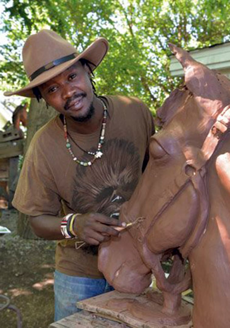 Man wearing a wide-brimmed hat carves a large clay horse statue outdoors, wearing a brown shirt and beaded necklace.
