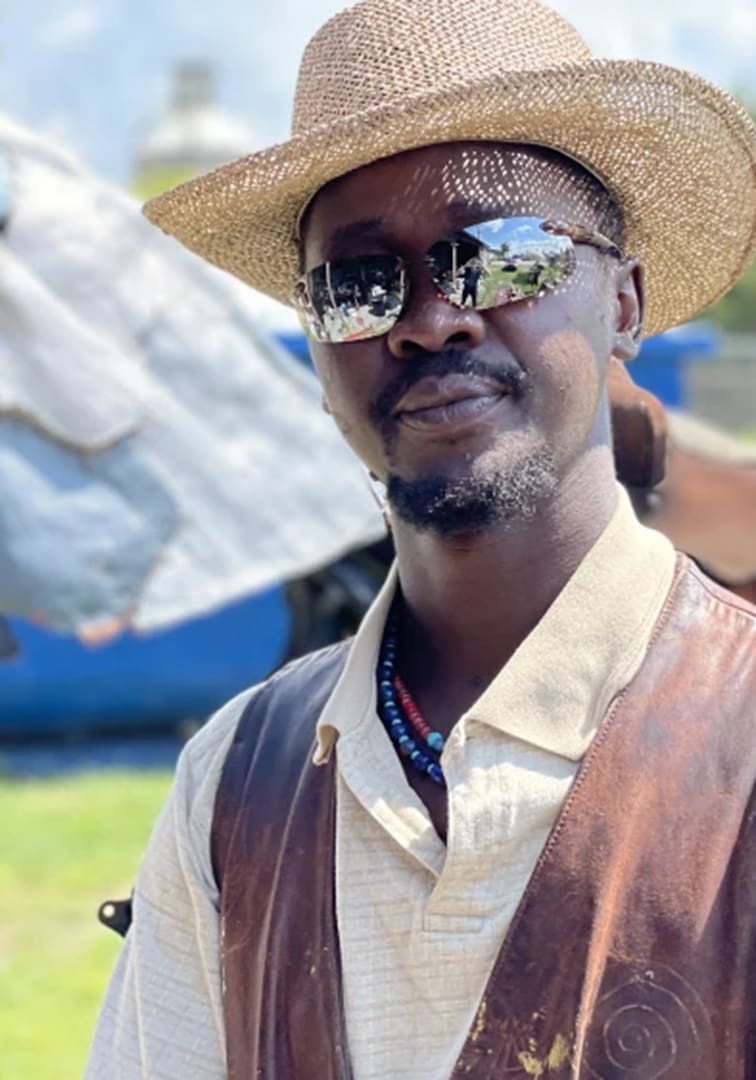 Man wearing a straw hat and mirrored sunglasses outdoors, with a cream shirt and brown leather vest.