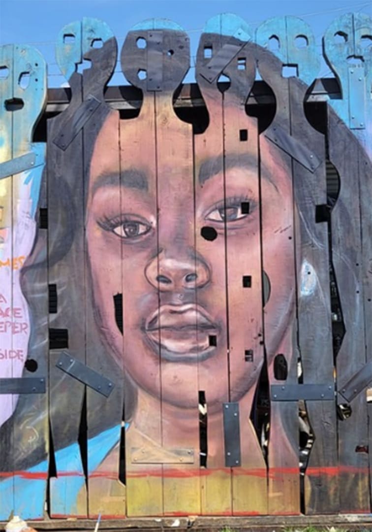 Portrait of a young Black boy painted on weathered vertical wooden slats forming a fence-like mural.