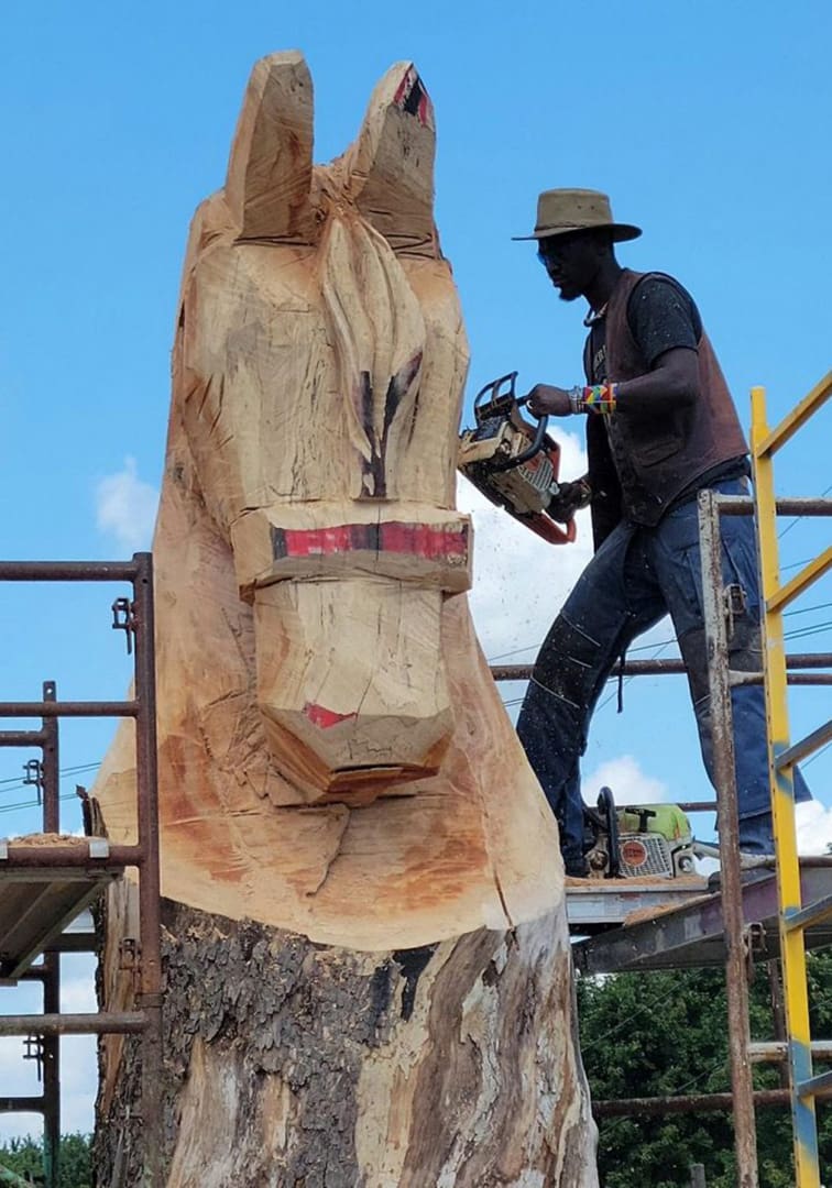 Carver with hat using a chainsaw to sculpt a large horse head from a tree trunk on a scaffold against a blue sky