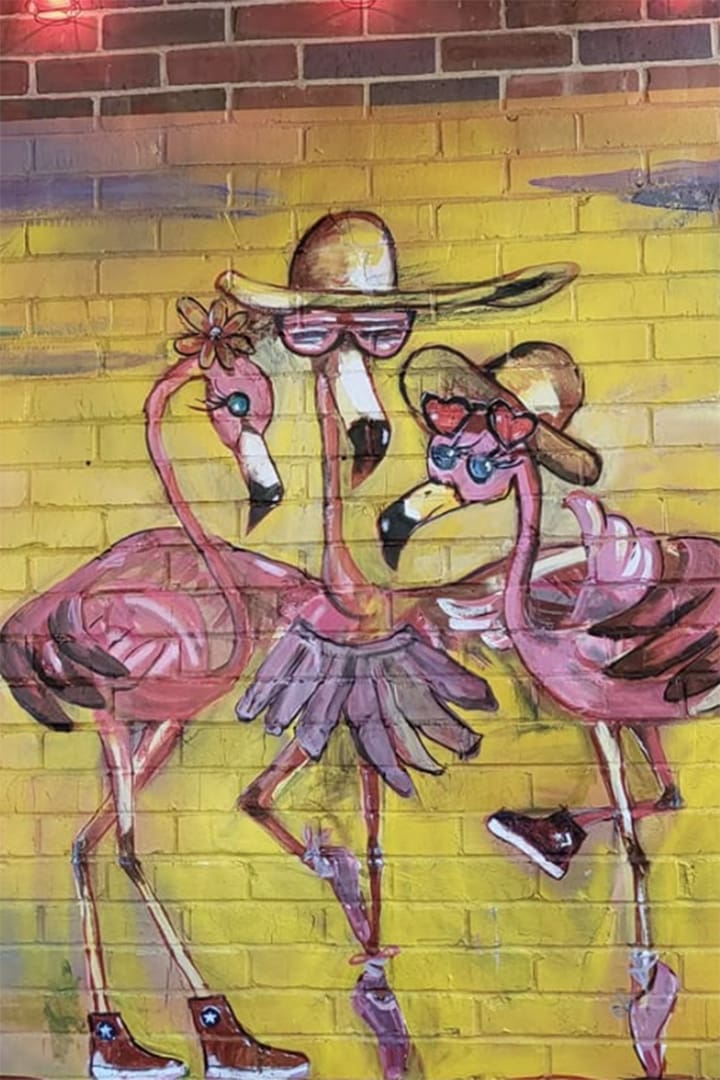 Two pink flamingos wearing hats and sunglasses stand together in a bright yellow graffiti mural on a brick wall.
