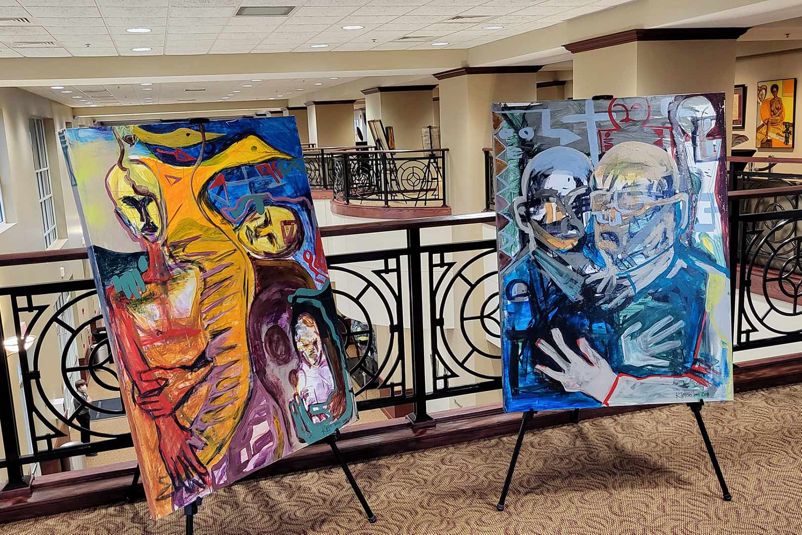 Two large abstract paintings on display in a well-lit indoor gallery, featuring vibrant colors and intertwined figures on easels by a decorative railing.