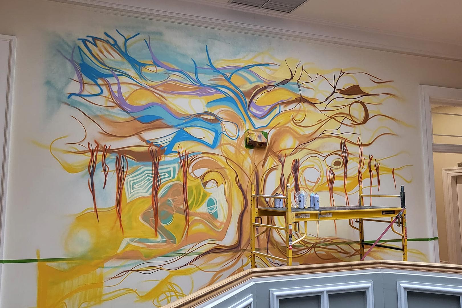 Large abstract mural with swirling blue, yellow, and orange lines on a wall, scaffold in front for painting.