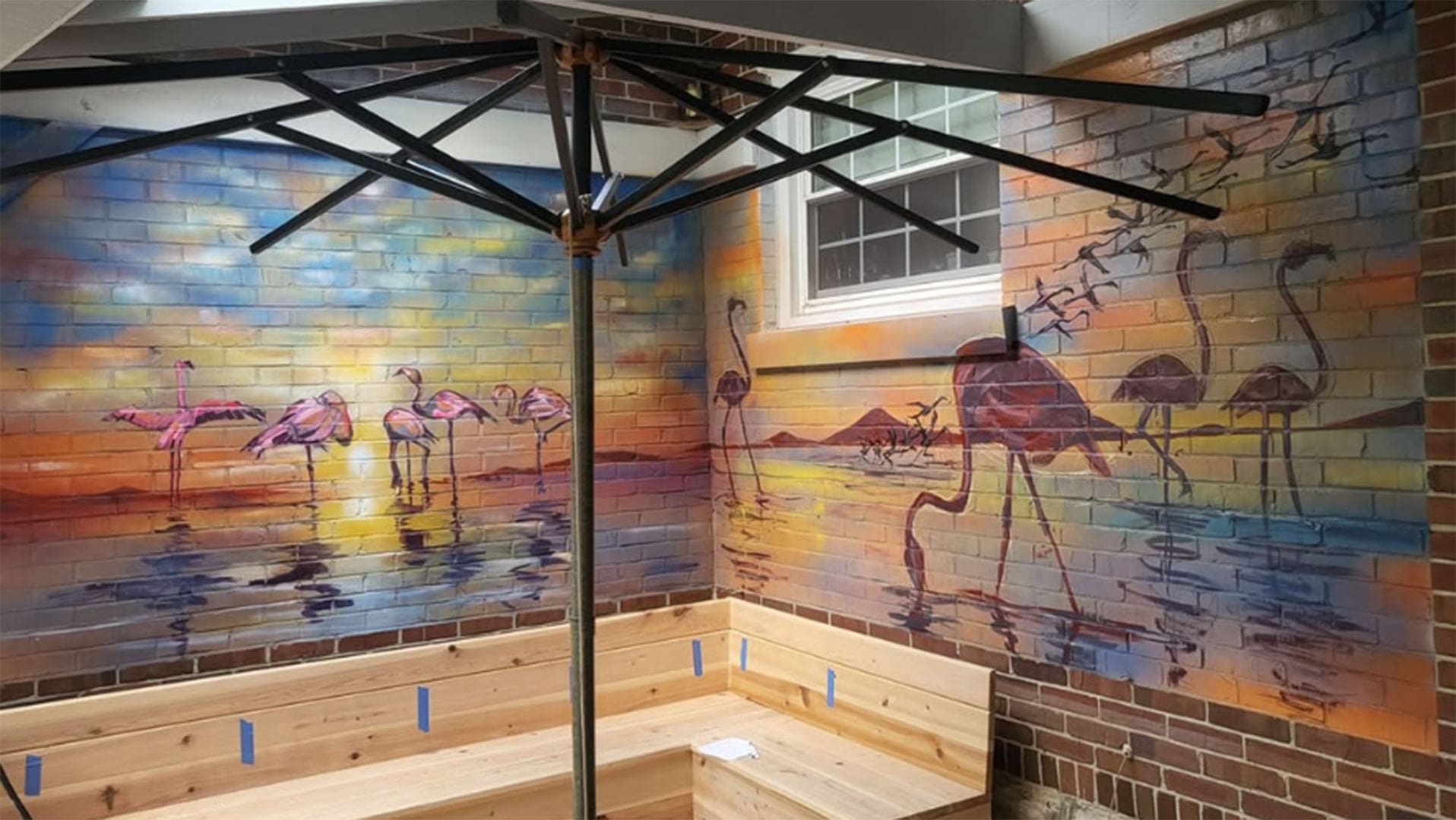 Brick-walled room featuring a sunset-flamingo mural along two walls, with a wooden bench and a metal umbrella frame overhead.