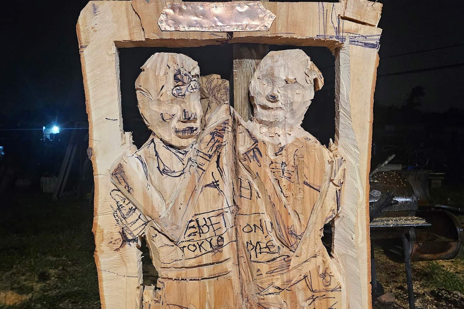 Crude wooden relief of two people carved in a rectangular frame, outdoors at night.