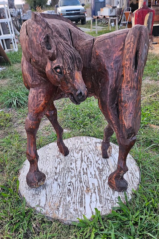 Wooden sculpture of two horses nuzzling heads, mounted on a round, weathered base outdoors in a grassy area.
