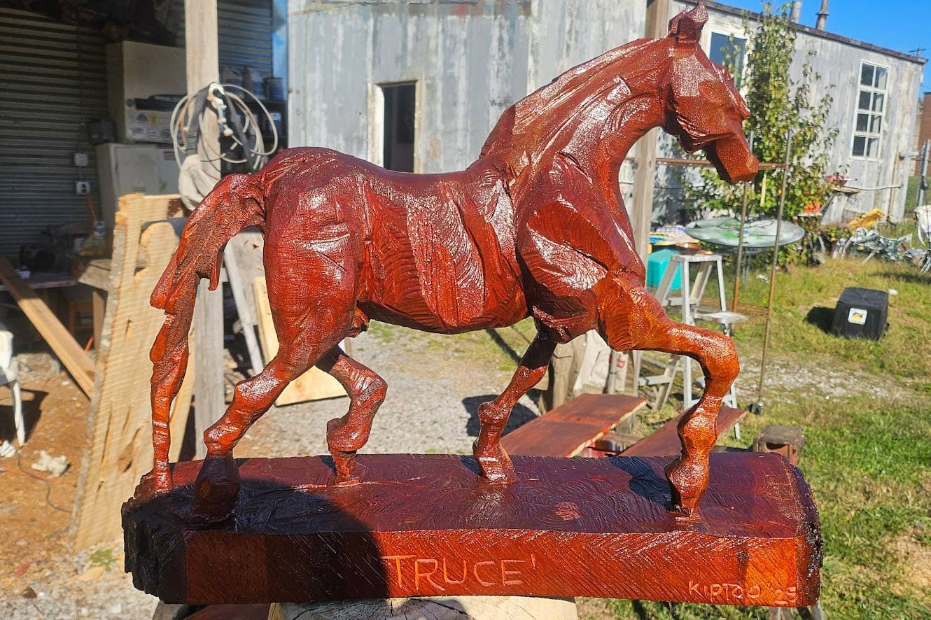 Red wooden horse sculpture on a rectangular pedestal outdoors in a workshop yard, base inscribed 'TRUCE'.