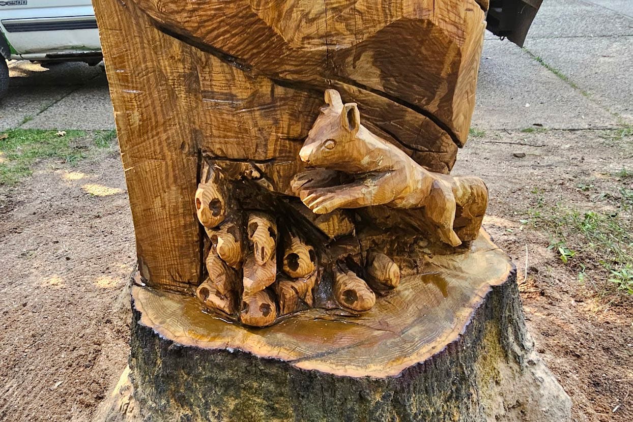 Wooden sculpture carved from a tree stump, a hand reaches toward a cluster of hollow log forms at the base of the carving.