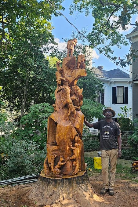 Tall carved wooden sculpture stacked with multiple figures, set in a yard beside a man in a hat for scale, against trees and a house background.