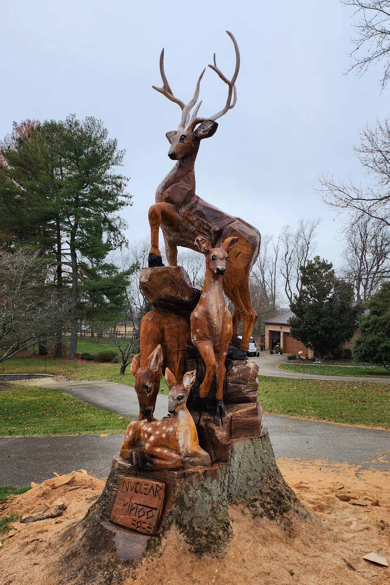 Wooden sculpture of a standing stag with large antlers on a rock pedestal, flanked by two smaller deer, outdoors in a yard. A carved plaque is mounted at the base.