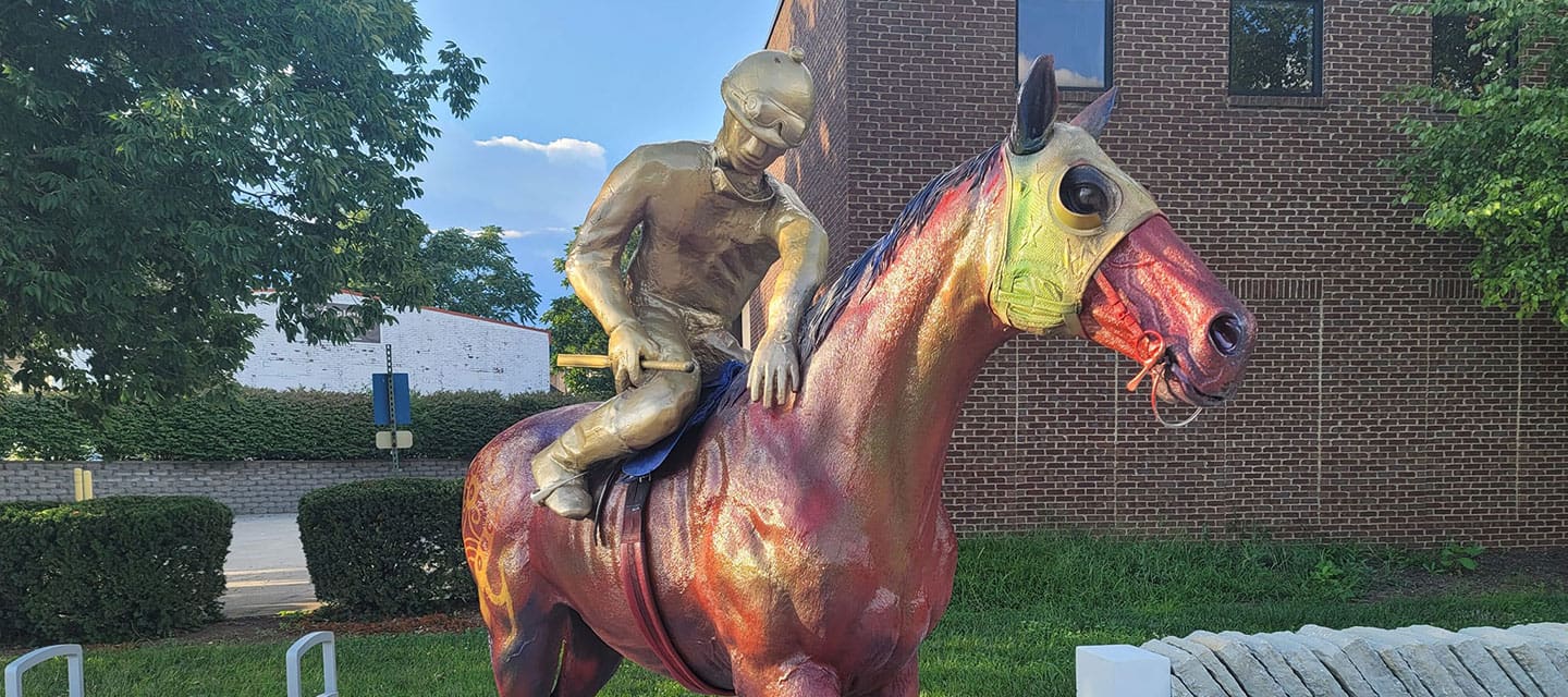 Colorful metallic statue of a jockey riding a horse outside a brick building.