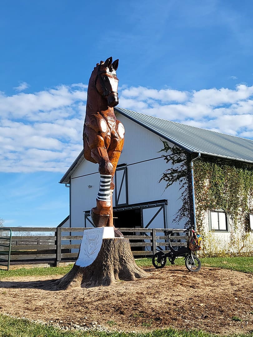 Large carved wooden horse statue mounted on a tree stump in a rural yard with barn and fence in the background.