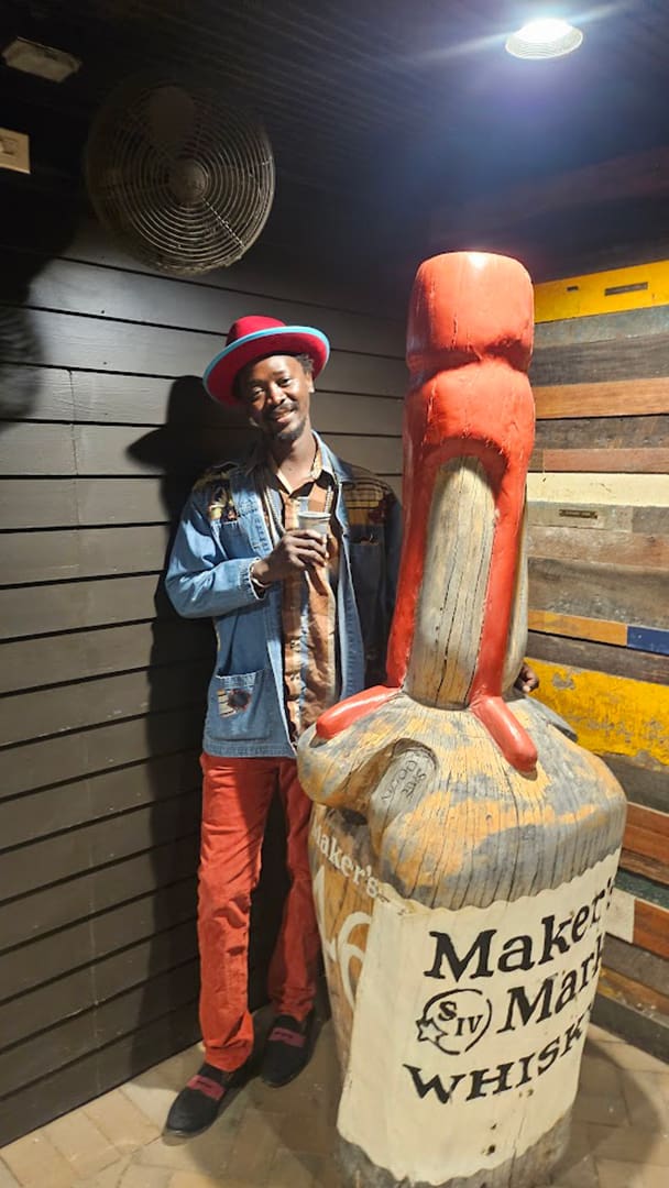 Smiling man in a red-brimmed hat and denim jacket posing beside a giant Maker's Mark whiskey bottle prop in a rustic room with wooden walls and a ceiling fan.