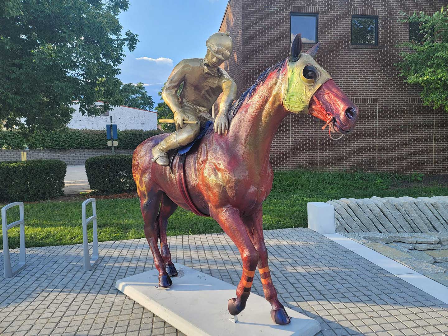 Rainbow-colored horse statue with a golden jockey, mounted on a white pedestal in an outdoor plaza beside a brick building.