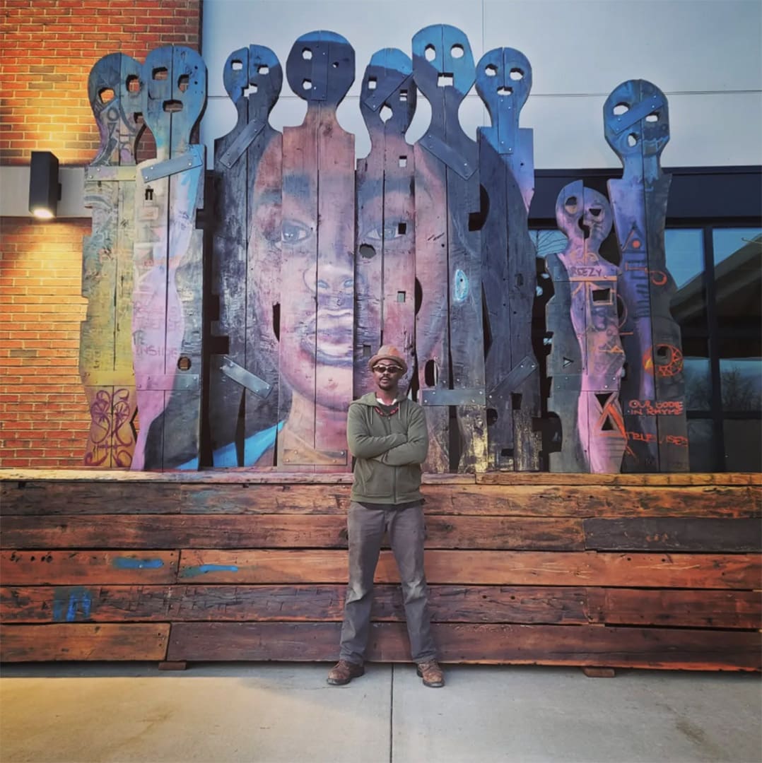 Man with crossed arms in a green jacket and hat stands in front of a tall wooden-panel mural with blue and purple figures.