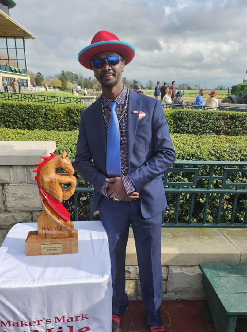 Man in a blue suit and bright red hat poses beside a wooden trophy on a table outdoors at a racetrack event.