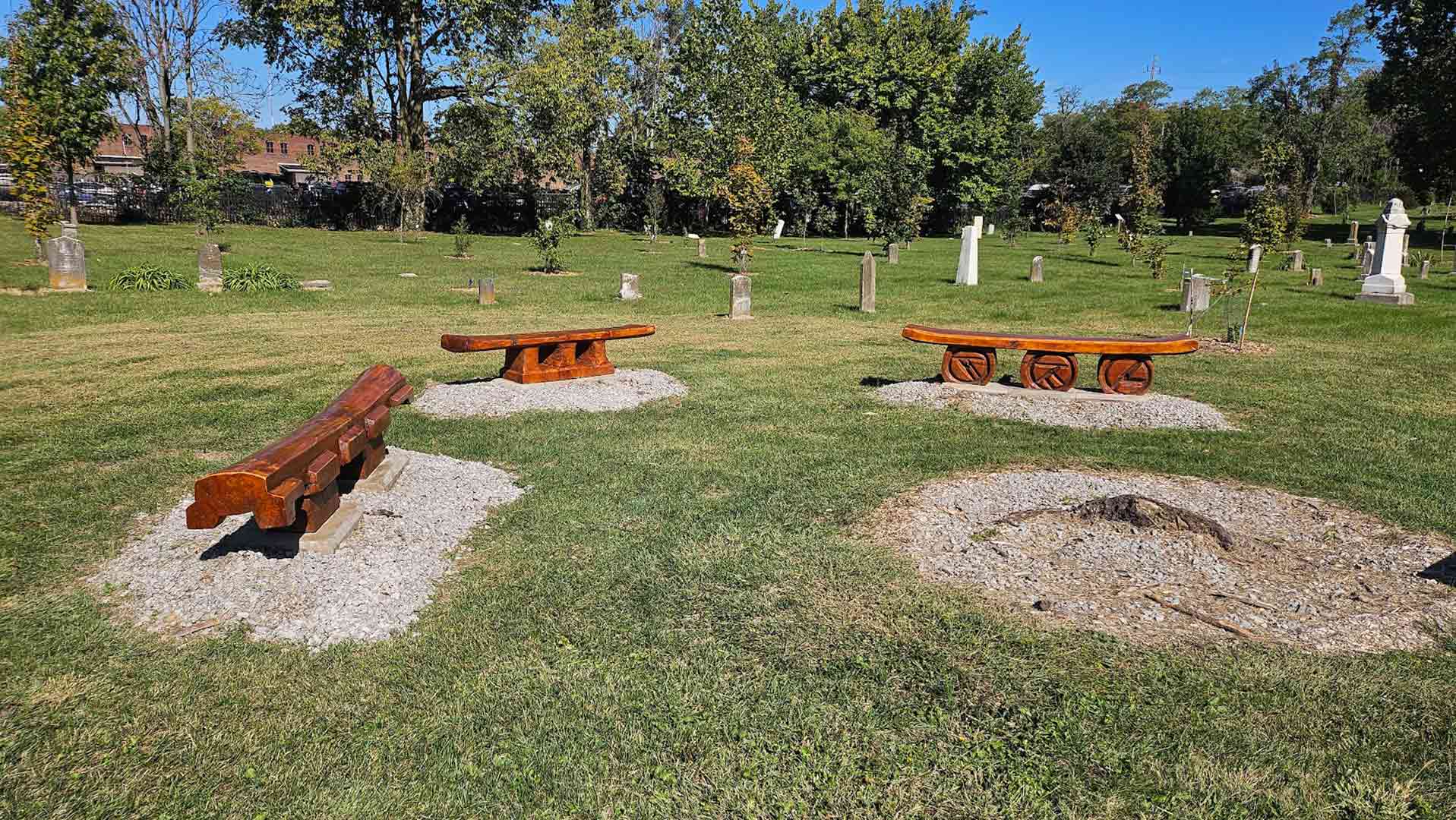 Wooden park benches on gravel pads in a sunny cemetery with headstones and trees in the background.