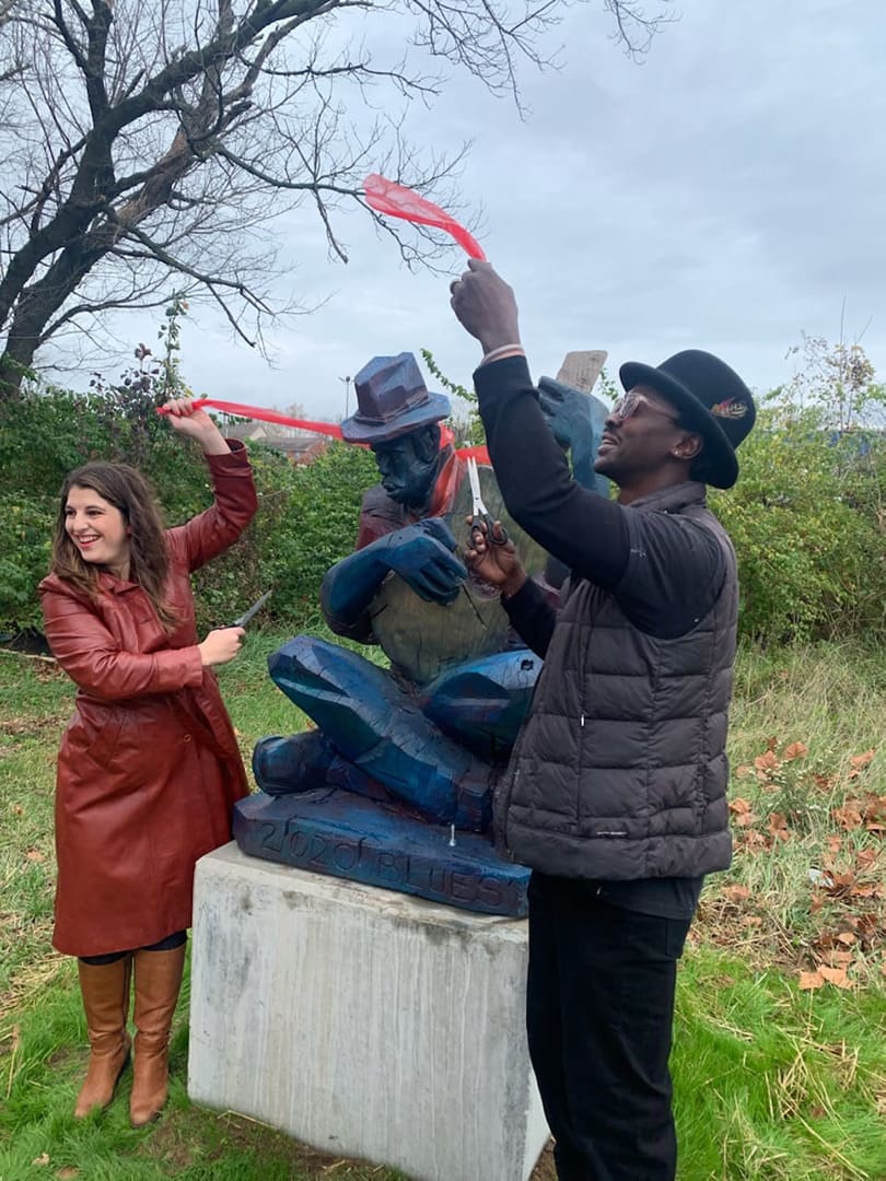 Two people cut a red ribbon in front of a blue seated sculpture outdoors.