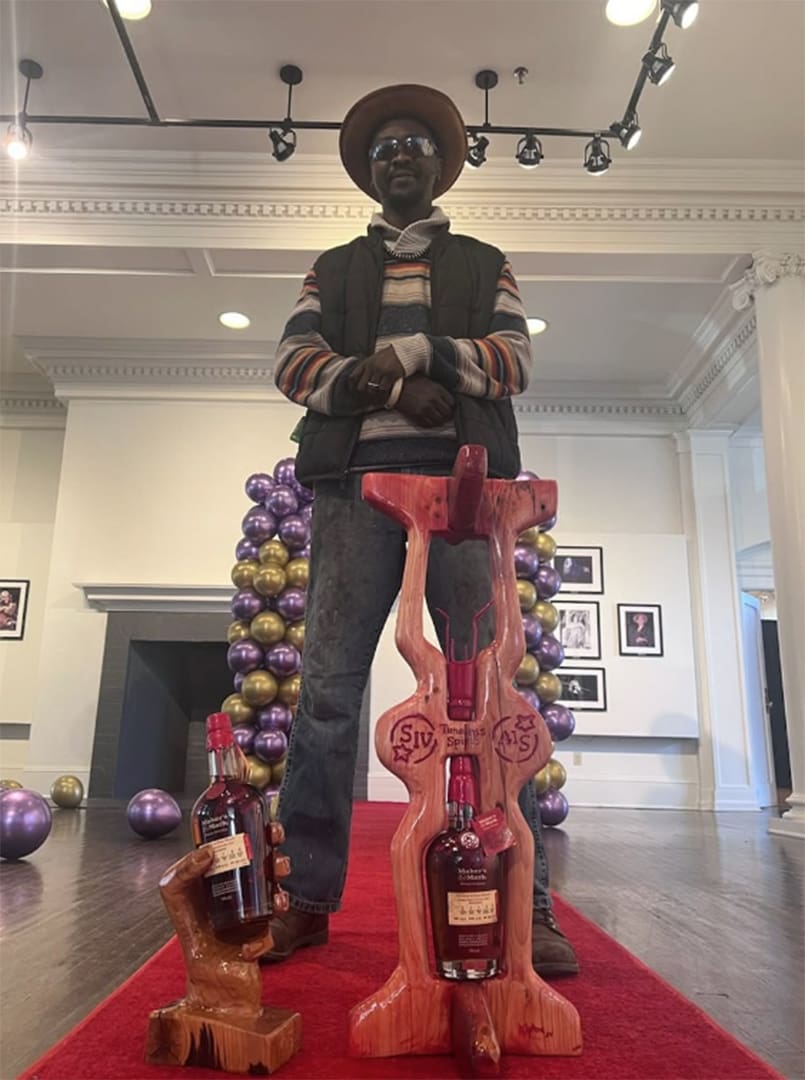 Man wearing a brown hat and striped sweater poses behind two carved wooden liquor bottle holders on a red carpet in a gallery.",
