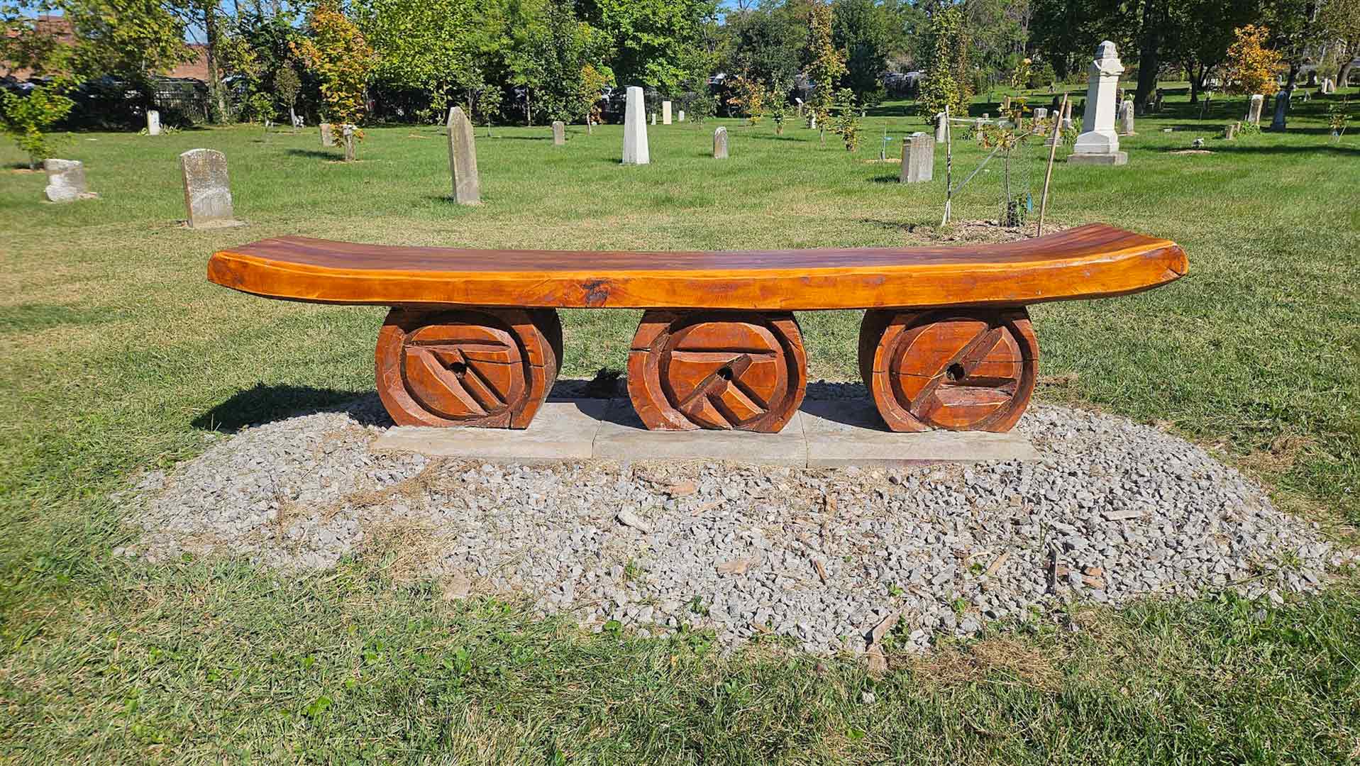 Curved wooden bench with three circular carved supports on a small gravel pedestal in a grassy cemetery park.