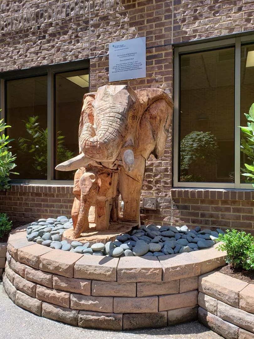 Wooden elephant fountain carved from logs, set in a circular stone planter in front of a brick building with windows.