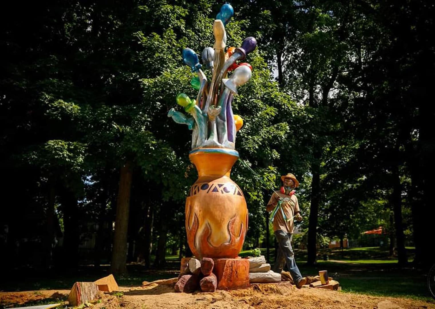 A gardener or worker stands beside a tall, colorful sculpture of a vase with multicolored bulbs, in a park setting under trees.