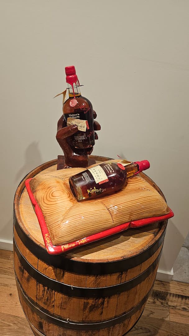 Whiskey display: a carved hand holds a Maker's Mark bottle above a wooden barrel, with another bottle resting on a wooden board atop the barrel.