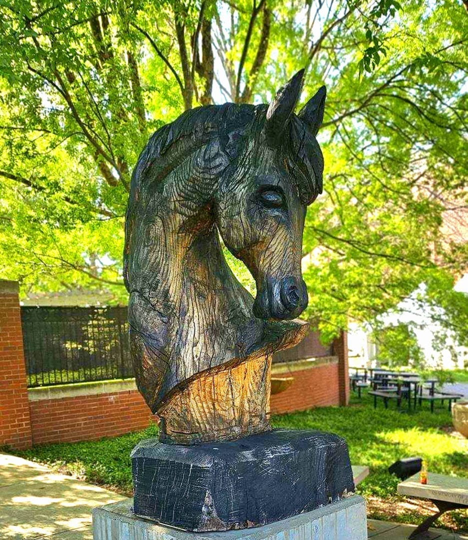 Bronze horse head sculpture on a pedestal outdoors with green trees and brick walls in the background.